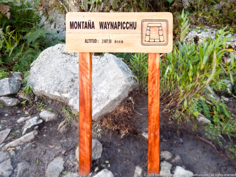 Wooden Montaña Waynapicchu sign with elevation information and a stair symbol beside rocks and vegetation.