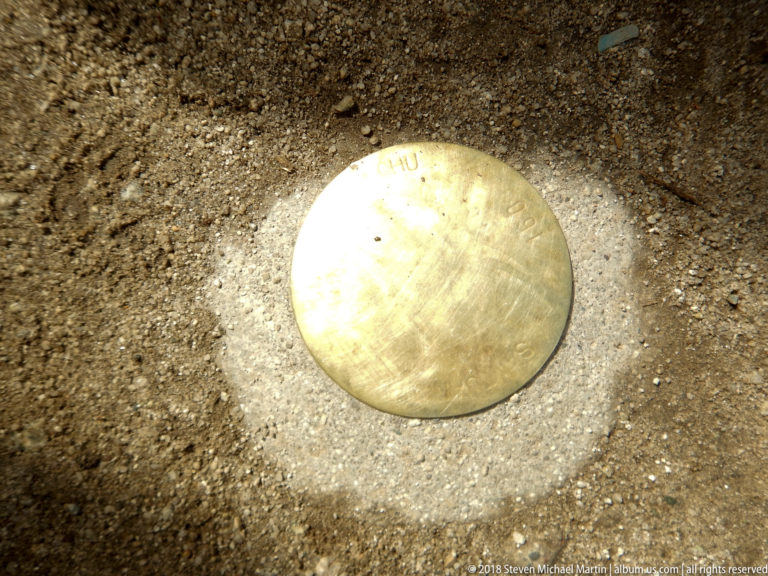 Circular metal survey marker set into compacted ground, with faint stamped lettering on its reflective surface.