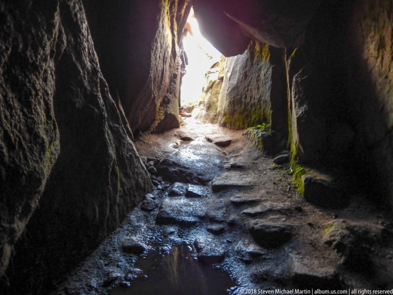 Dark narrow rock passage with a damp stone floor and daylight at the far opening on the Huayna Picchu route.