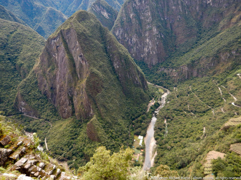 Deep green valley below Machu Picchu with a winding river and switchback road beneath steep rock faces.