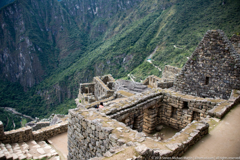 Overhead view of roofless stone rooms and a surviving gabled wall at Machu Picchu above a steep valley.