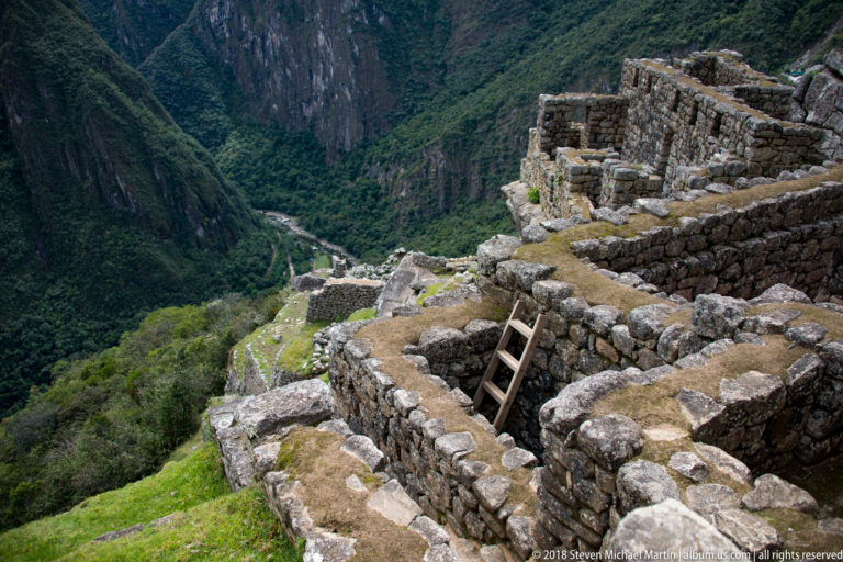 Roofless stone rooms at Machu Picchu overlook a deep green valley with a river far below.