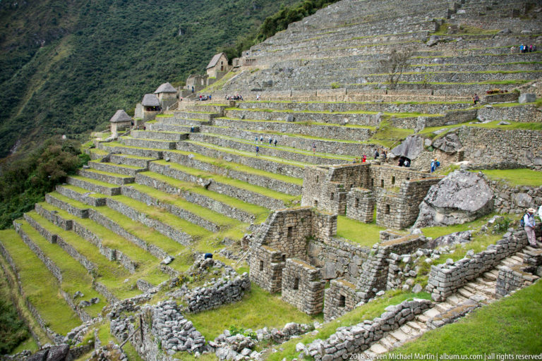 Parallel stone agricultural terraces descend a steep green slope with small masonry structures at Machu Picchu.