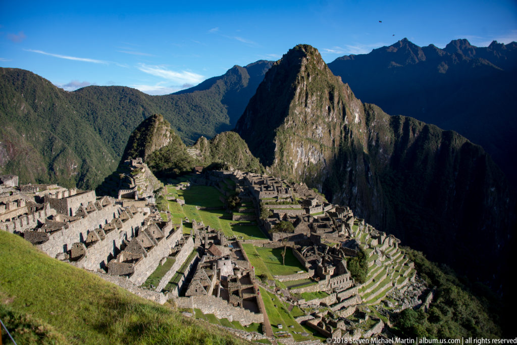 Elevated view across Machu Picchu’s roofless masonry, green lawns, terraces, and Huayna Picchu.