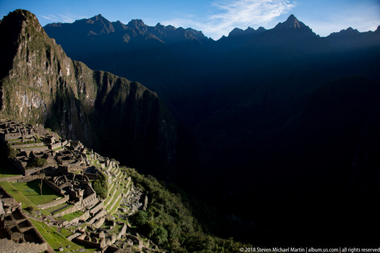 Oblique view of Machu Picchu’s terraces and stone buildings beneath steep shadowed Andean ridges.