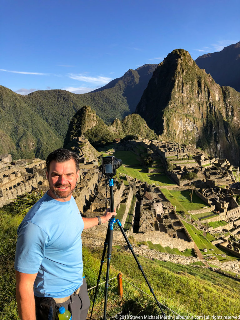 Visitor in a blue shirt beside a camera on a tripod with Machu Picchu and Huayna Picchu behind.