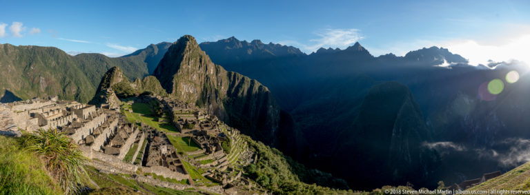 Backlit Machu Picchu and Huayna Picchu with deep mountain shadows, low sun, and visible lens flare.