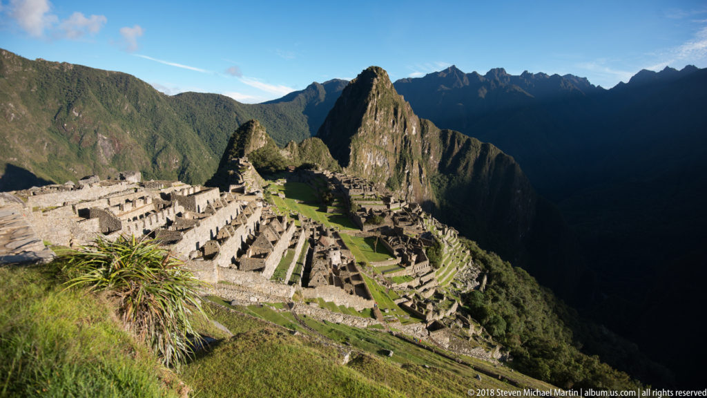Foreground grasses frame Machu Picchu’s stone buildings, green terraces, and Huayna Picchu.