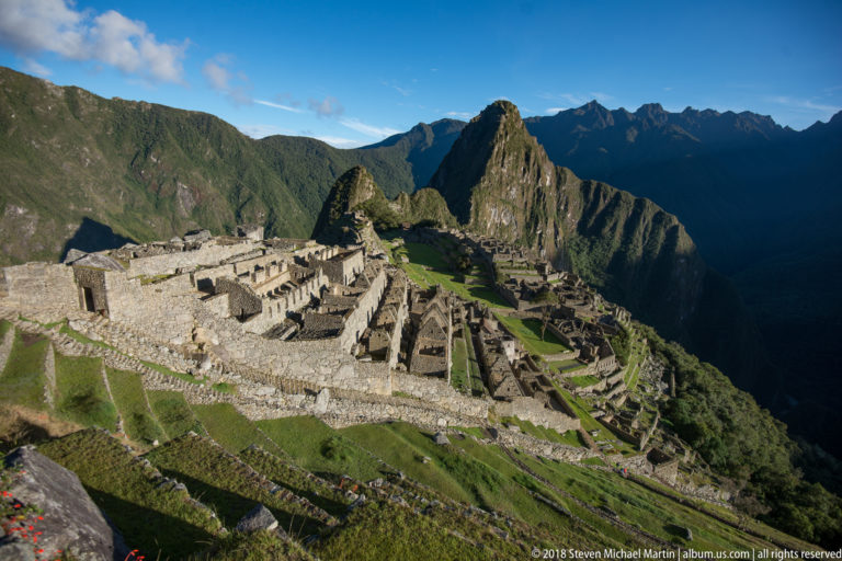 Wide late-day panorama of Machu Picchu beneath Huayna Picchu and shadowed Andean ridges.