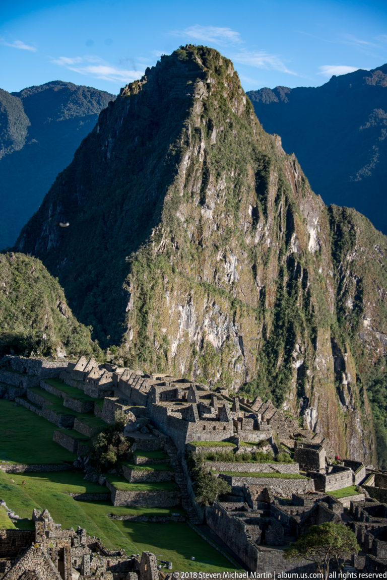 Stone compounds and green lawns line the Machu Picchu ridge beneath a rounded mountain.