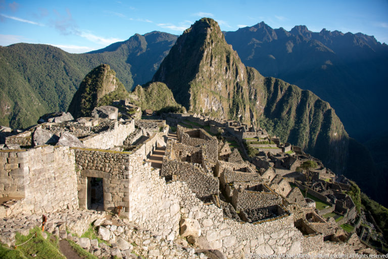 Roofless stone rooms and passages descend across Machu Picchu with Huayna Picchu behind.