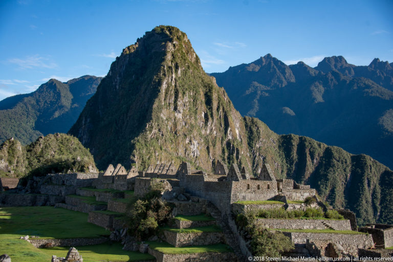 Huayna Picchu rises above green lawns, terraces, and roofless stone buildings at Machu Picchu.