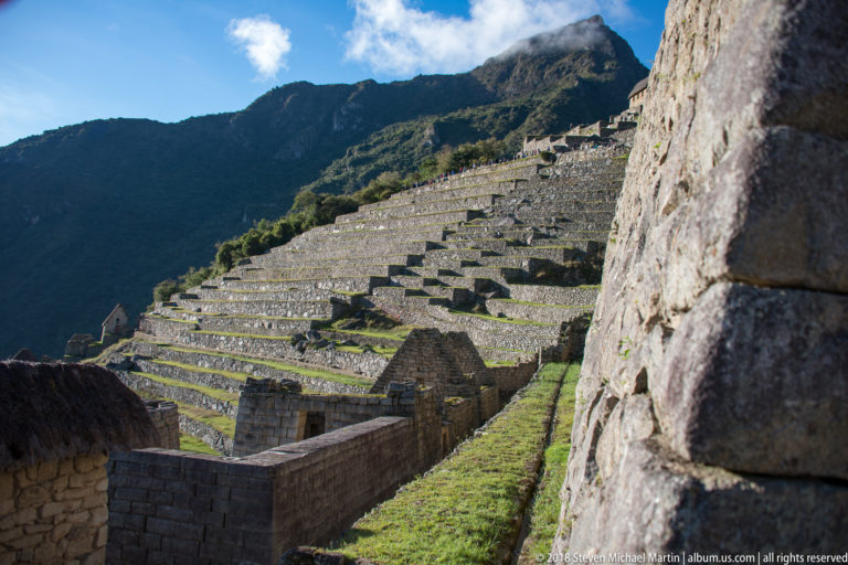 Sunlit terraces rise beyond a narrow passage between dark stone walls at Machu Picchu.