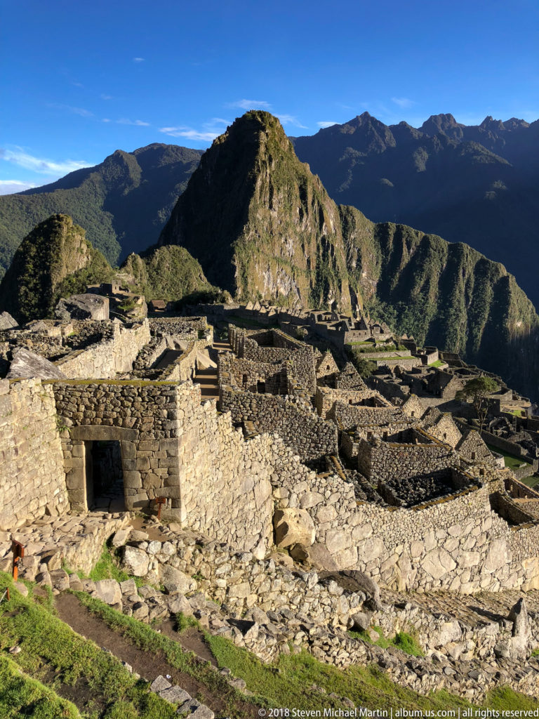 Broad view of roofless Machu Picchu stone rooms and terraces beneath a rounded green mountain.