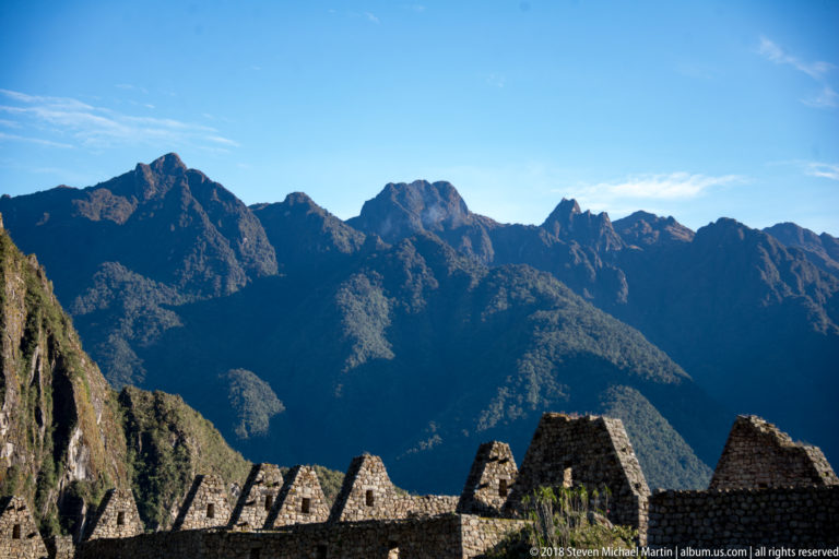 Jagged Andean peaks rise beyond silhouetted roofless stone gables at Machu Picchu.
