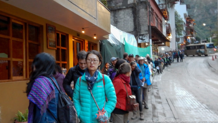 Long line of visitors with backpacks waiting along a narrow street in Machu Picchu Pueblo.