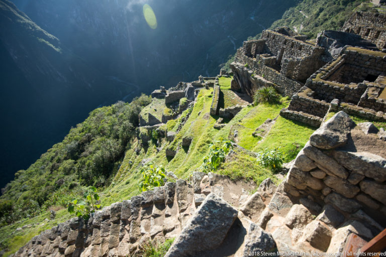 Sunlit stone terraces and low masonry descend steeply toward a dark mountain valley at Machu Picchu.