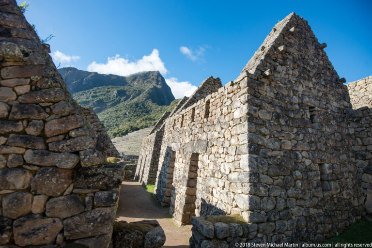 Roofless stone buildings with tall gabled walls and narrow doorways along a passage at Machu Picchu.