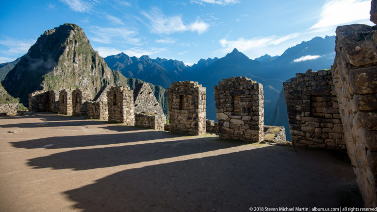 Roofless stone buildings with gabled walls cast long shadows along a path, with Huayna Picchu behind.