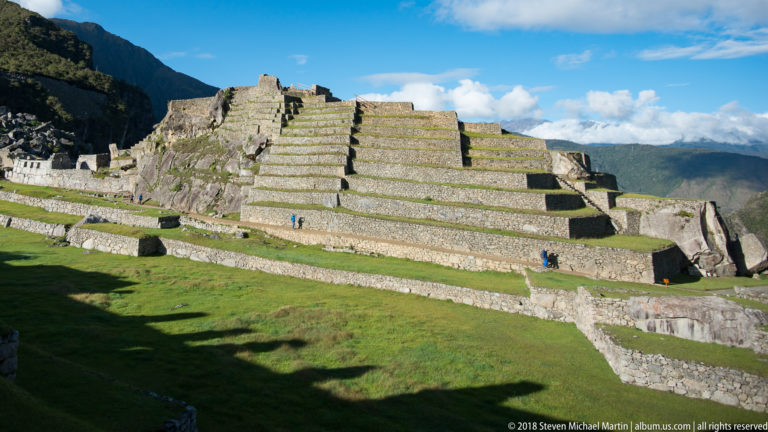 Large stepped stone terraces rise above a broad green lawn with a few visitors at Machu Picchu.