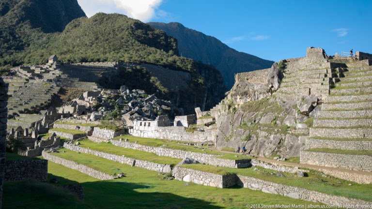Layered green terraces, stone retaining walls, roofless buildings, and large boulders at Machu Picchu.