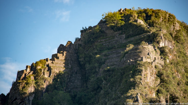Steep Huayna Picchu peak with stone terraces and small masonry structures near its sunlit summit.