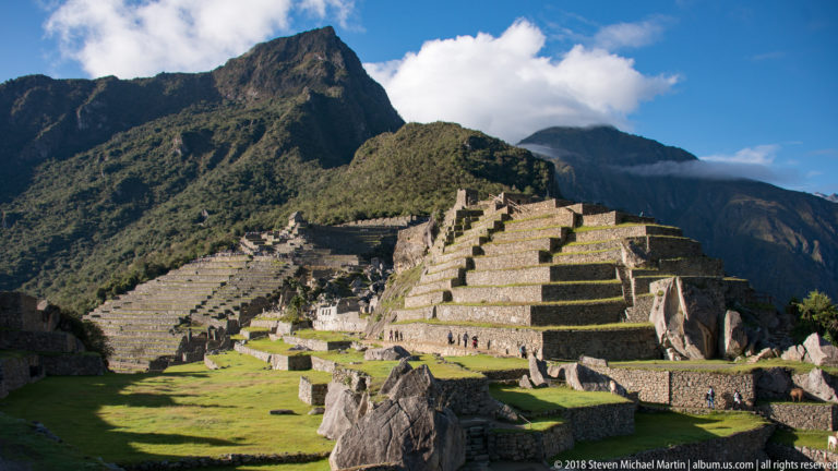 Large stepped stone terraces and lower masonry groups at Machu Picchu beneath green mountains and clouds.