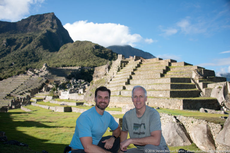 Two men posing before stepped stone terraces and roofless masonry at Machu Picchu.