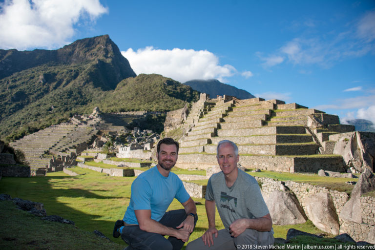 Two men posing on a green lawn before large stepped stone terraces and masonry at Machu Picchu.