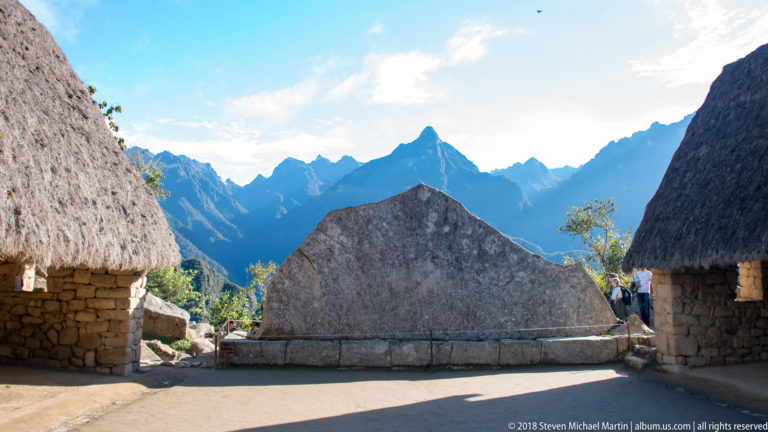 Large angular stone centered between thatched buildings with jagged Andean peaks beyond at Machu Picchu.