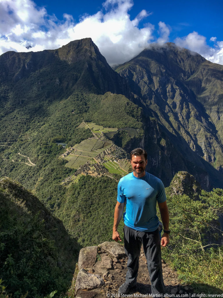 Man walking uphill on a rocky trail with Machu Picchu and mountain ridges behind him.