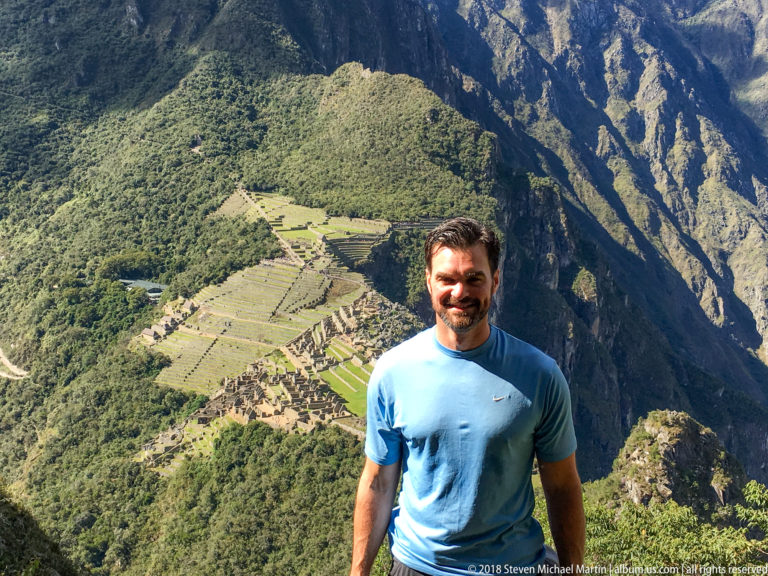 Man in a blue shirt standing above Machu Picchu with terraces and stone buildings visible below.