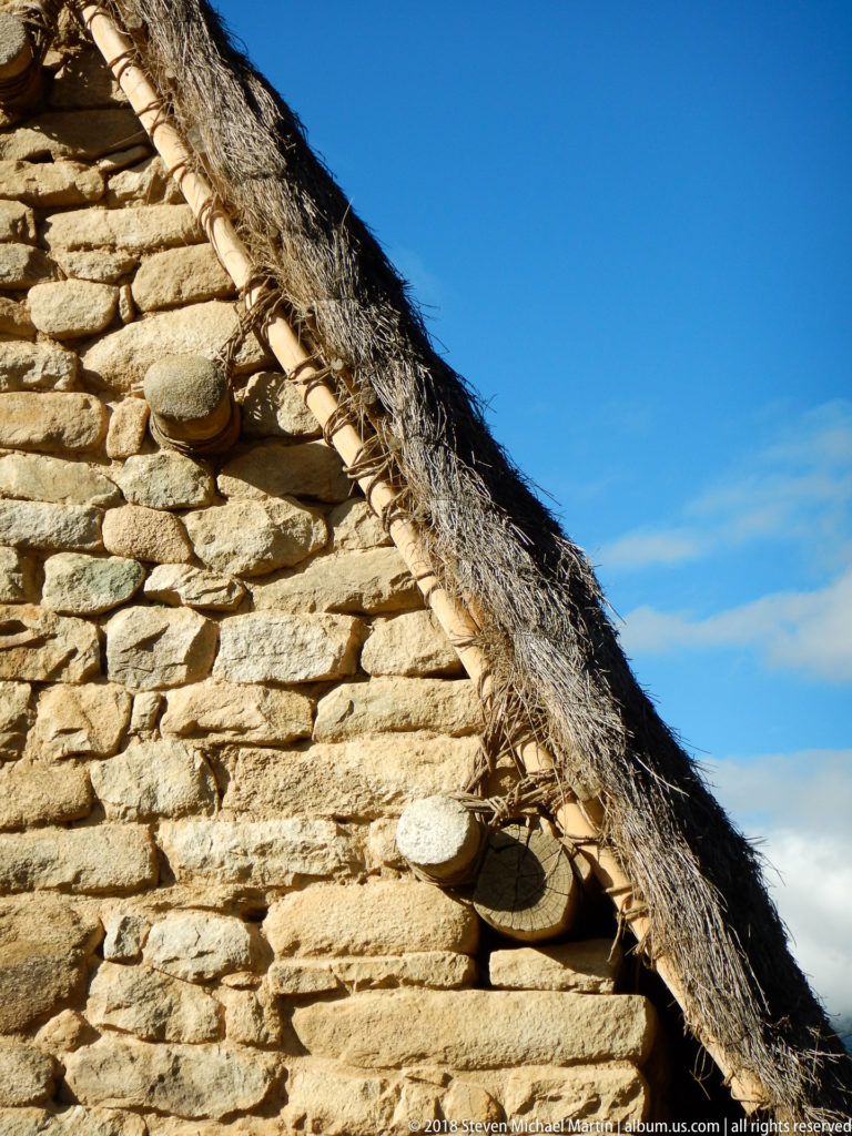 Stone gable wall with a dark reconstructed thatched roof against a bright blue sky at Machu Picchu.