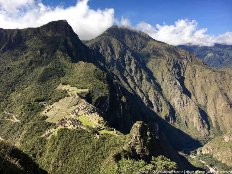 Sunlit Machu Picchu ridge beneath immense Andean mountain walls and bright clouds.