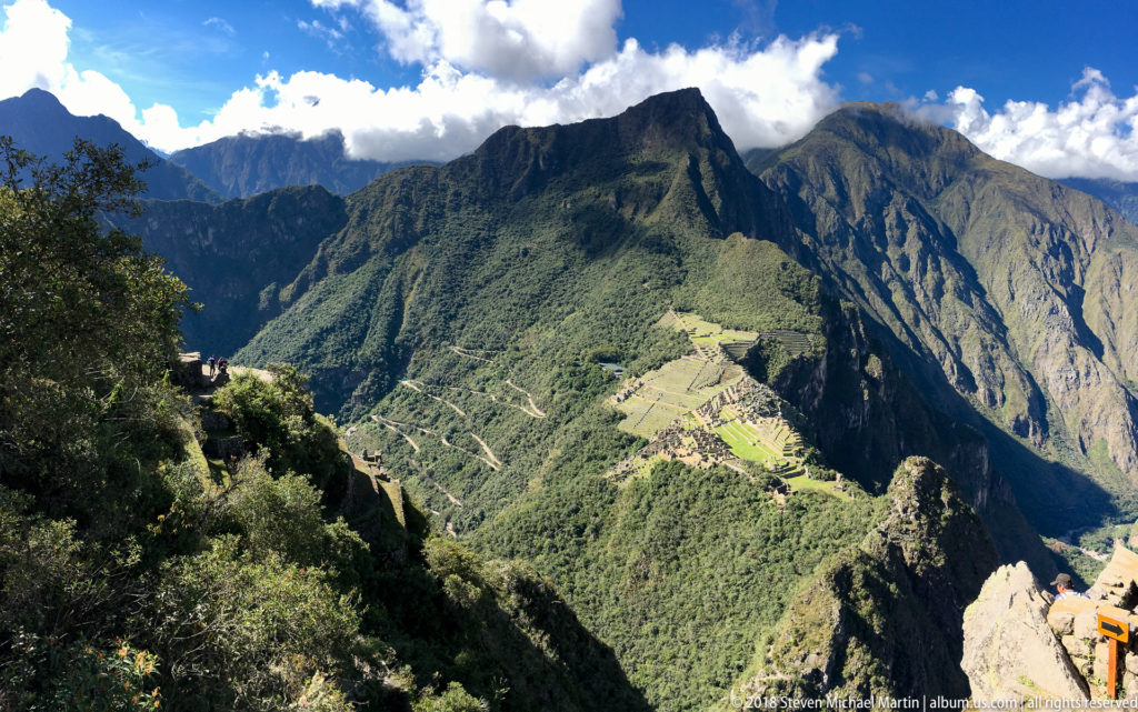 High view from Huayna Picchu toward Machu Picchu on a green ridge surrounded by steep mountains.