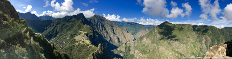 Jagged green and rock-faced Andean peaks rise beneath bright white clouds near Machu Picchu.