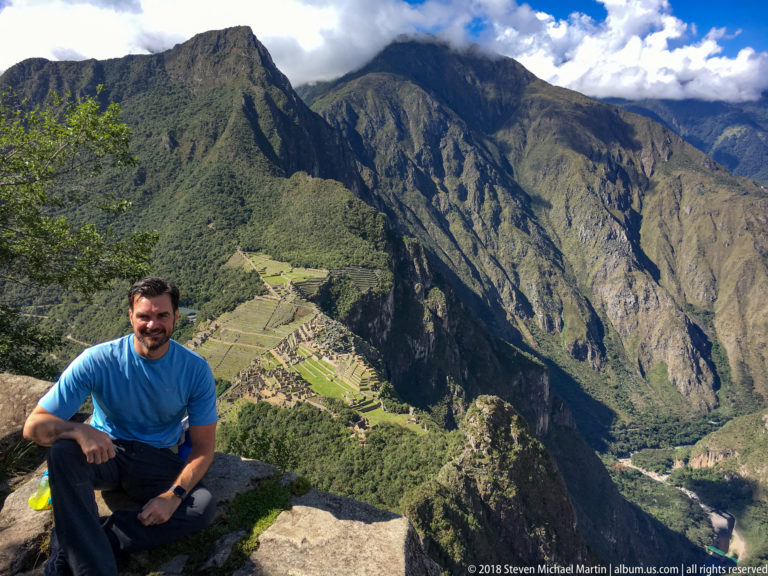 Seated man on a stone ledge above Machu Picchu with steep Andean mountains filling the background.