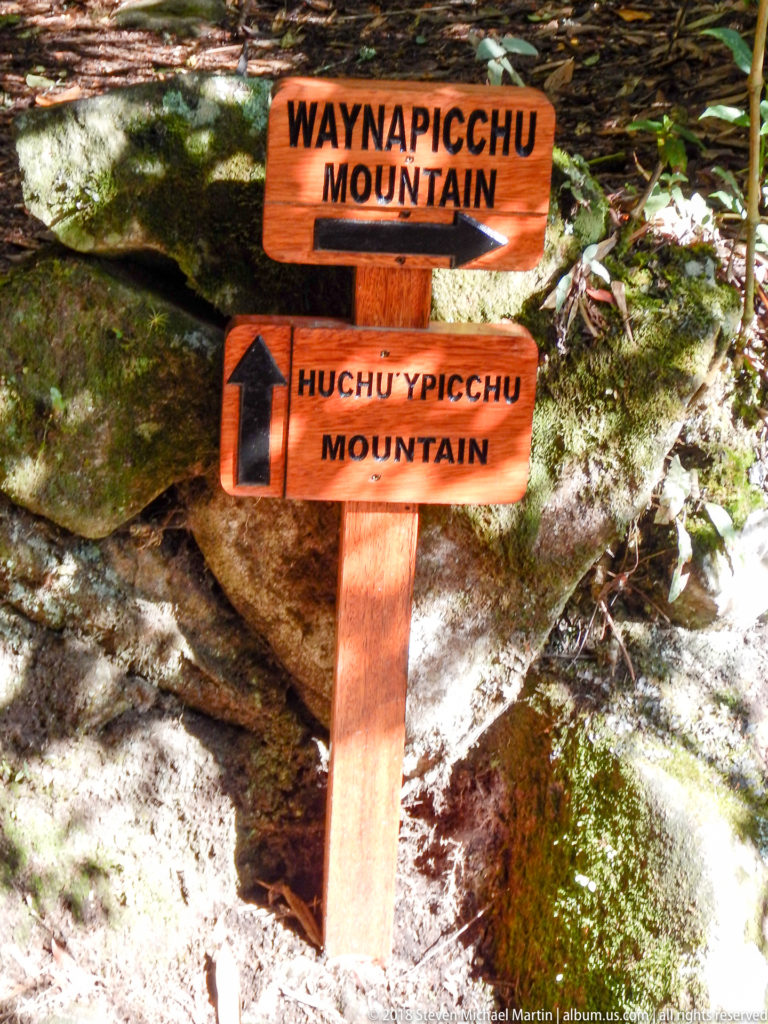 Wooden junction sign pointing toward Waynapicchu Mountain and Huchuypicchu Mountain among rocks and vegetation.