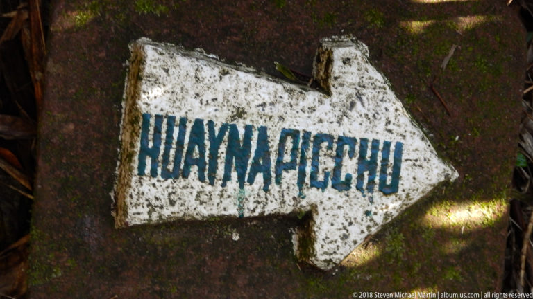 White painted arrow labeled HUAYNA PICCHU on a dark stone surface beside the mountain trail.