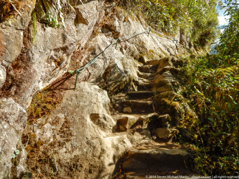 Steep irregular stone steps climb beside exposed bedrock and a fixed handline on the Huayna Picchu trail.