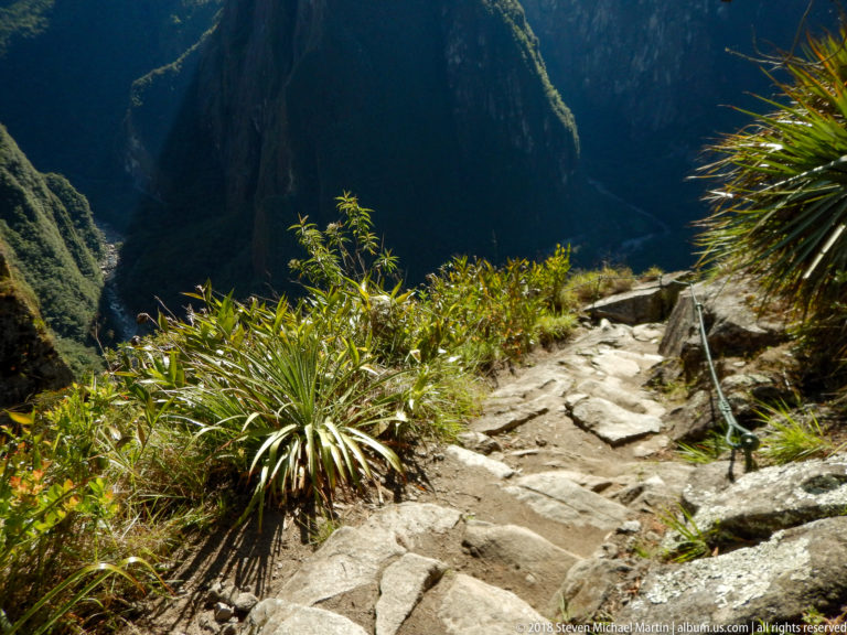 Steep stone steps and a metal handline descend past spiky plants toward a shadowed mountain valley.