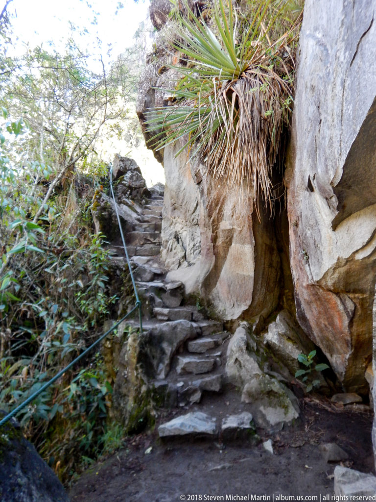 Narrow stone stair with a metal handrail passing between large rocks and dense plants on Huayna Picchu.