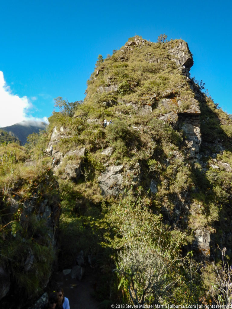 Sunlit rocky Huayna Picchu outcrop with low shrubs and grasses beneath a clear blue sky.