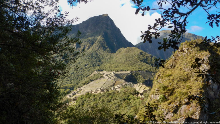 Machu Picchu’s terraces and buildings seen through shaded foliage beneath the steep peak of Huayna Picchu.