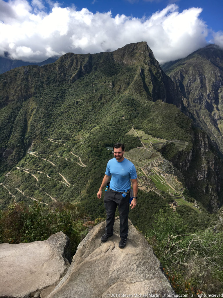 Man stepping across an exposed rock ledge on Huayna Picchu with Machu Picchu visible behind.
