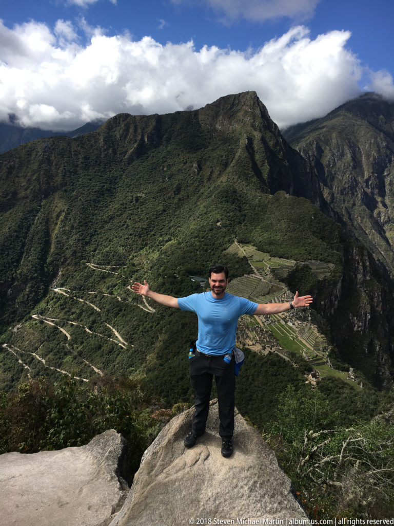 Visitor standing with arms outstretched on a Huayna Picchu rock ledge above Machu Picchu.