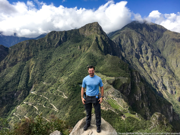 Man in a blue shirt standing on a rock ledge with Machu Picchu and green mountains far below.
