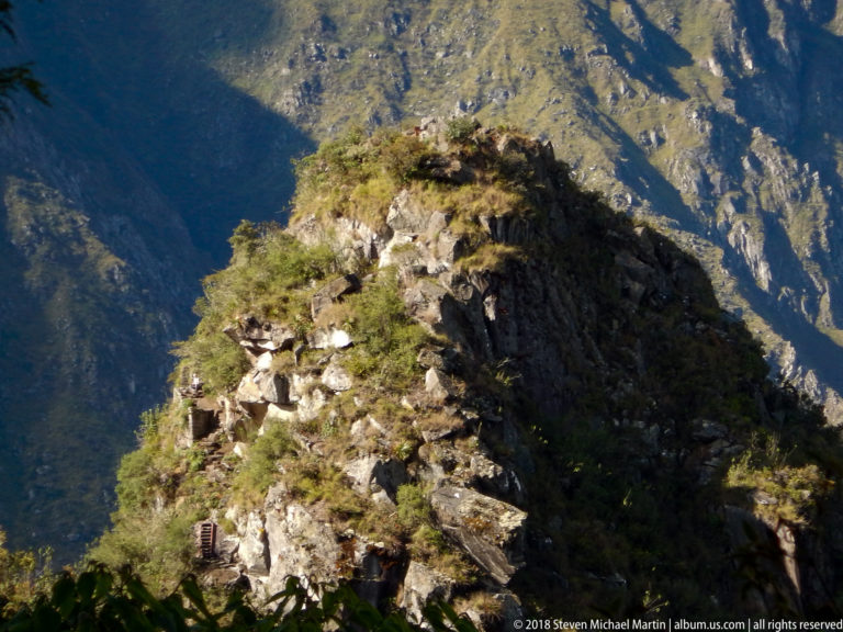 Rocky Huayna Picchu ridge with grasses and small trail elements above a deep mountain valley.