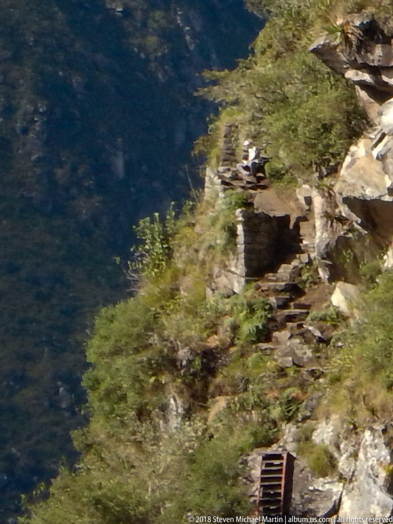 Distant stonework and wooden trail elements cling to a steep vegetated slope above a dark valley.