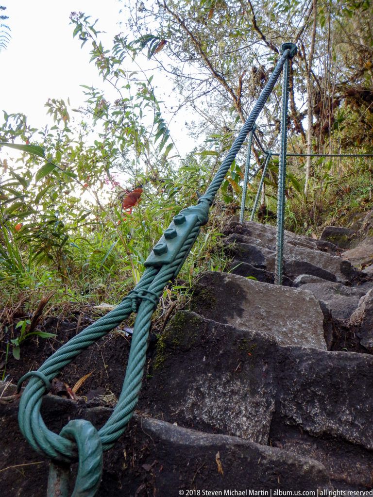 Thick green rope handline tied to a metal post beside steep stone steps and vegetation on Huayna Picchu.